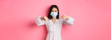 Coronavirus, sosyal uzaklık ve yaşam tarzı konsepti. Yüz maskesi takmış hayal kırıklığına uğramış Asyalı kadın yargılayıcı görünüyor, baş parmaklarını göstermiyor ve katılmıyor, pembe arka planda duruyor..