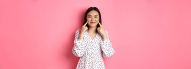 Tatlı, utangaç Asyalı kız yanaklarını dürtüyor, kameraya mutlu bir şekilde gülümsüyor, romantik beyaz elbiseler giyiyor, pembe arka planda duruyor..