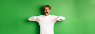 Şaşırmış ve heyecanlı genç kızıl saçlı adam sevinç ve şaşkınlık çığlıkları atıyor, muhteşem tanıtımlara bakıyor, parmakları aşağıda, yeşil arka planda duruyor..