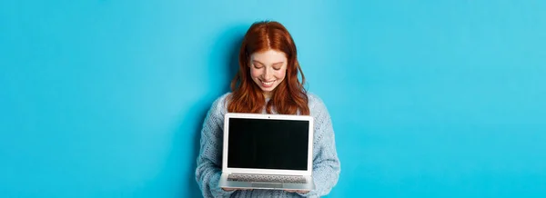 Cute redhead woman in sweater, showing and looking at laptop screen with pleased smile, demonstrating something online, standing over blue background.
