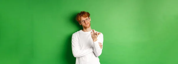 Young man with red messy hair and glasses, smiling pleased and pointing hand at camera, your turn gesture, standing over green background.