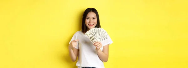 Young asian woman smiling, showing prize money, pointing finger at dollars, standing over yellow background.