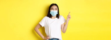 Covid, sağlık ve salgın hastalık konsepti. Tıbbi maskeli ve beyaz tişörtlü Asyalı kadın sol üst köşedeki logoyu işaret ediyor..