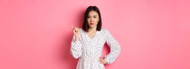 Üzgün ve kasvetli Asyalı kız kaşlarını çatmış, sağ üst köşeyi işaret ediyor ve mutsuz bir şekilde kameraya bakıyor, pembe arka planda duruyor..