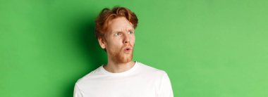 Kafası karışmış kızıl saçlı adam sola bakıyor ve endişeli görünüyor, yeşil arka plana karşı duruyor..