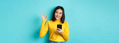 E-ticaret ve online alışveriş konsepti. Asyalı kadın portresi tamam işareti gösteriyor ve cep telefonu kullanıyor, övgü uygulaması kullanıyor, mavi arka planda duruyor..