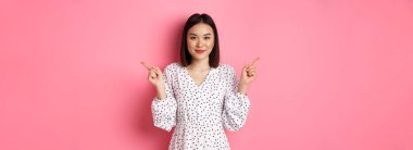 Güzel Asyalı kadın alışverişte seçim yapıyor, parmaklarını yana doğru gösteriyor ve değişkenleri gösteriyor, kameraya gülümsüyor, pembe arka planda duruyor..