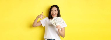 Gülümseyen Asyalı kadın parayı işaret ediyor, dolar gösteriyor, sarı arka planda duruyor..