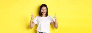 Beyaz tişörtlü, güzel Asyalı bir kadın, baş parmağını kaldırıyor ve gülümsüyor, iyi bir teklif sunuyor, ürün öneriyor, sarı arka planda tatmin olmuş bir şekilde duruyor..