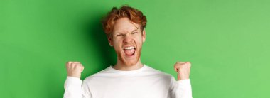 Şanslı genç adamın yakın çekimi, zaferi kutluyor, sevinçle evet diye bağırıyor ve yumruk sallıyor, yeşil arka planda zafer kazanıyor..