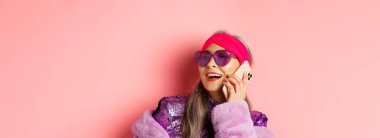 Moda. Güneş gözlüklü, disko kıyafetli, telefonla konuşan, gülümseyen ve pembe arka planda duran son sınıf Asyalı son sınıf öğrencisinin yakın çekimi..