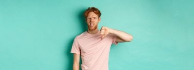 Hayal kırıklığına uğramış kızıl saçlı adam baş parmağını ve dilini gösteriyor. Sevmiyor ve onaylamıyor. Turkuaz arka planda duruyor..