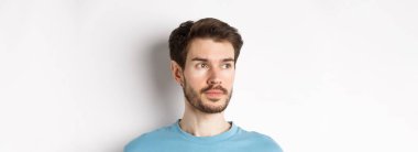 Sakallı ve bıyıklı yakışıklı erkek modelin yakın çekimi, düşünceli bir yüzle sola bakıyor, beyaz arka planda duruyor..