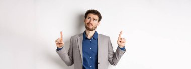 Şüpheci ve tereddütlü takım elbiseli genç işadamı hayal kırıklığına uğramış bir sırıtışla yukarı bakıyor, anlaşmadan emin değil, beyaz arka planda tatminsiz duruyor..