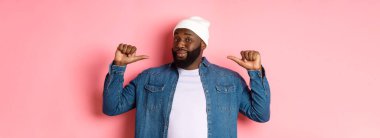 Kendinden emin ve küstah siyah adam kendini işaret ediyor, kameraya bakıyor kendinden emin, gösteriş meraklısı, pembe arka planda duruyor..