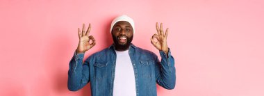 Hipster bereli tatmin olmuş Afro-Amerikan bir adam, onaylama işaretleri gösteriyor, iyi bir teklifi övüyor ve evet diyor, mutlu gülümsüyor, pembe arka planda duruyor..