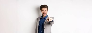 Sensin. Takım elbiseli, gülümseyen yakışıklı yönetici kamerayı işaret ediyor, birini seçiyor ve mutlu görünüyor, beyaz arka planda duruyor..