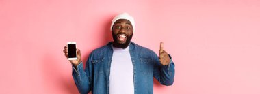 Çevrimiçi alışveriş ve teknoloji kavramı. Mutlu Afro-Amerikan hipster adam baş parmağını kaldırıyor ve mobil ekran, uygulamayı övüyor, pembe arka planda duruyor..