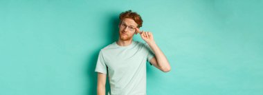 Şüpheci kızıl saçlı bir adam aptal ya da deli birini azarlıyor, parmağıyla kafasını işaret ediyor ve kameraya bakıyor, arka planda duruyor..