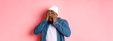 Meraklı siyah adam gözlerini kapatıyor ama parmaklarından bakıyor, kameraya şaşkın şaşkın bakıyor, pembe arka planda hippi beresi takıyor..