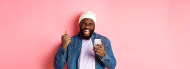 Teknoloji ve online alışveriş konsepti. Mutlu siyah adam seviniyor, uygulamayı kazanıyor, akıllı telefonu tutuyor ve evet diye bağırıyor, pembe arka planda duruyor..