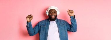 Mutlu genç Afro-Amerikalı adam kulaklıkla dans ediyor ve müzik dinliyor, yumruklarını sallıyor ve gülümsüyor, pembe arka planda duruyor..