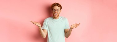 Şaşırmış gözlüklü kızıl saçlı adamın görüntüsü, ellerini yana doğru açıyor ve etkilenmiş görünüyor, pembe arka planda duruyor..