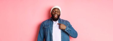Hipster bereli şaşkın siyah adam kendini işaret ediyor, kameraya şaşkın şaşkın bakıyor, pembe arka planda duruyor..