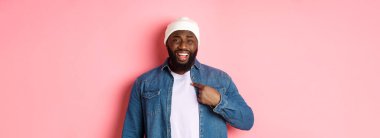 Gücenmiş ve kafası karışmış Afro-Amerikan bir adam kendini işaret ediyor, kameraya bakıyor, rahatsız oluyor, suçlanıyor, pembe arka planda duruyor..