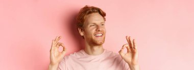 Tişörtlü, gülümseyen, tatmin olmuş ve sola bakan, onay veren, harika ürünü öven, pembe arka planda duran yakışıklı kızıl saçlı adamı yakından çek..