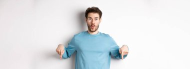 Sakallı, sıradan mavi tişörtlü, hayret verici ve aşağıdan işaret eden genç adam, şuraya bak, beyaz arka planda duruyor..