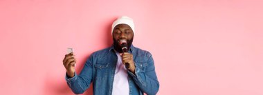 Yakışıklı Afro-Amerikalı adam karaoke söylüyor, akıllı telefon uygulamasında şarkı sözleri okuyor ve mikrofon tutuyor, pembe arka planda duruyor..
