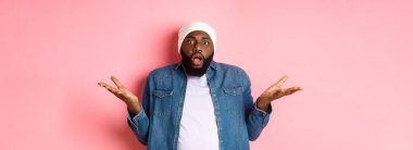 Kafası karışmış ve şok olmuş Afro-Amerikan erkeği ellerini yana açtı ve yere düştü, kameraya hayranlık ve şaşkınlıkla baktı, pembe arka planda hiçbir şey bilmeden durdu..