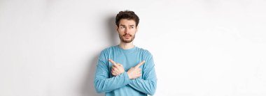 Kararsız adam seçim yapıyor, parmaklarını iki varyanta doğrultuyor ve karar veriyor, sol tarafta dalgın dalgın bakıyor, beyaz arka planda duruyor..