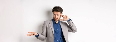 Delirdin mi sen? Takım elbiseli genç adam tuhaf ya da aptalca bir hatayı işaret ederek omuz silkiyor, beyaz arka planda duruyor..