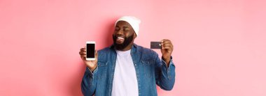 İnternetten alışveriş. Tatmin olmuş siyahi adam onaylamak için başını salladı, gülümsedi ve telefona baktı, kredi kartı ve akıllı telefon ekranını gösterdi, pembe arka planda durdu..