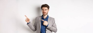 Takım elbiseli yakışıklı adam sağ üst köşedeki logoyu işaret ediyor. Şirketin afişini ve iddialı gülümsemesini gösteriyor. Beyaz arka planda duruyor..