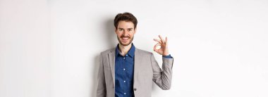 Gülümseyen başarılı takım elbiseli işadamı tamam işareti gösteriyor, tatmin olmuş görünüyor, beyaz arka plana karşı duruyor..