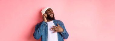 Mutlu Afro-Amerikalı adam cep telefonuyla konuşuyor, gülüyor ve gülümsüyor, pembe arka plan üzerinde bere ve kot gömlek ile duruyor..