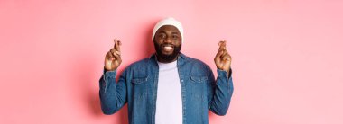 İyimser Afro-Amerikalı bir adam dilek diliyor, parmaklarını çapraz tutuyor ve gülümsüyor, pembe arka planda duruyor..