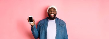 Bereli ve kot gömlekli yakışıklı hippi gülümsüyor, mutlu yüzlü cep telefonu ekranını gösteriyor, uygulama öneriyor, pembe arka planda duruyor..