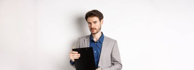 Genç erkek CEO elinde belgelerle pano tutuyor ve kameraya bakıyor, ofisi inceliyor, beyaz arka planda duruyor..