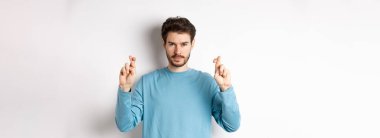 Umut dolu ve kendinden emin genç adam şans dilemek ya da dua etmek için parmaklarını çapraz yapıyor, sonuçları tahmin ediyor, beyaz arka planda duruyor..