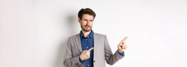 Takım elbiseli hoşnutsuz işadamı şüpheci bir sırıtışla sol tarafa bakıyor, pankarttan hoşlanmıyor, onaylanmayan promosyon teklifi, beyaz arka planda duruyor..