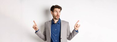 Takım elbiseli kararsız erkek girişimci parmaklarını yana doğru gösteriyor, iki varyant arasında seçim yapıyor, sol logoya dalgın bakıyor, beyaz arka planda duruyor..