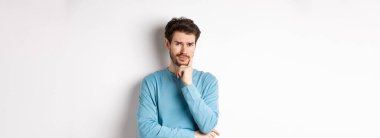 Şüpheci genç adam somurtuyor, endişeli düşünceli bakıyor, beyaz arka plan üzerinde üzgün duruyor.