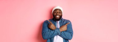 Mutlu genç Afro-Amerikalı hippi adam parmaklarını yana doğru gösteriyor, gülümsüyor ve iki seçenek gösteriyor, teklifleri gösteriyor, pembe arka planda duruyor..