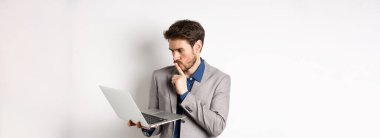 Düşünceli iş adamı dizüstü bilgisayarda çalışıyor ve düşünüyor, bilgisayar ekranında düşünceli görünüyor, beyaz arka planda takım elbise içinde duruyor..