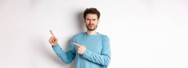 Hayal kırıklığına uğramış ve üzgün genç bir adam kötü bir teklif yapıyor, parmaklarını doğru gösteriyor ve suratını asıyor, beyaz arka planda şüpheci bir yüzle duruyor..