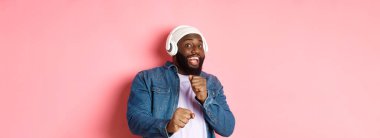 Mutlu Afro-Amerikalı adam dans ediyor, kulaklıkla müzik dinliyor ve pembe arka planda kameraya bakıyor..